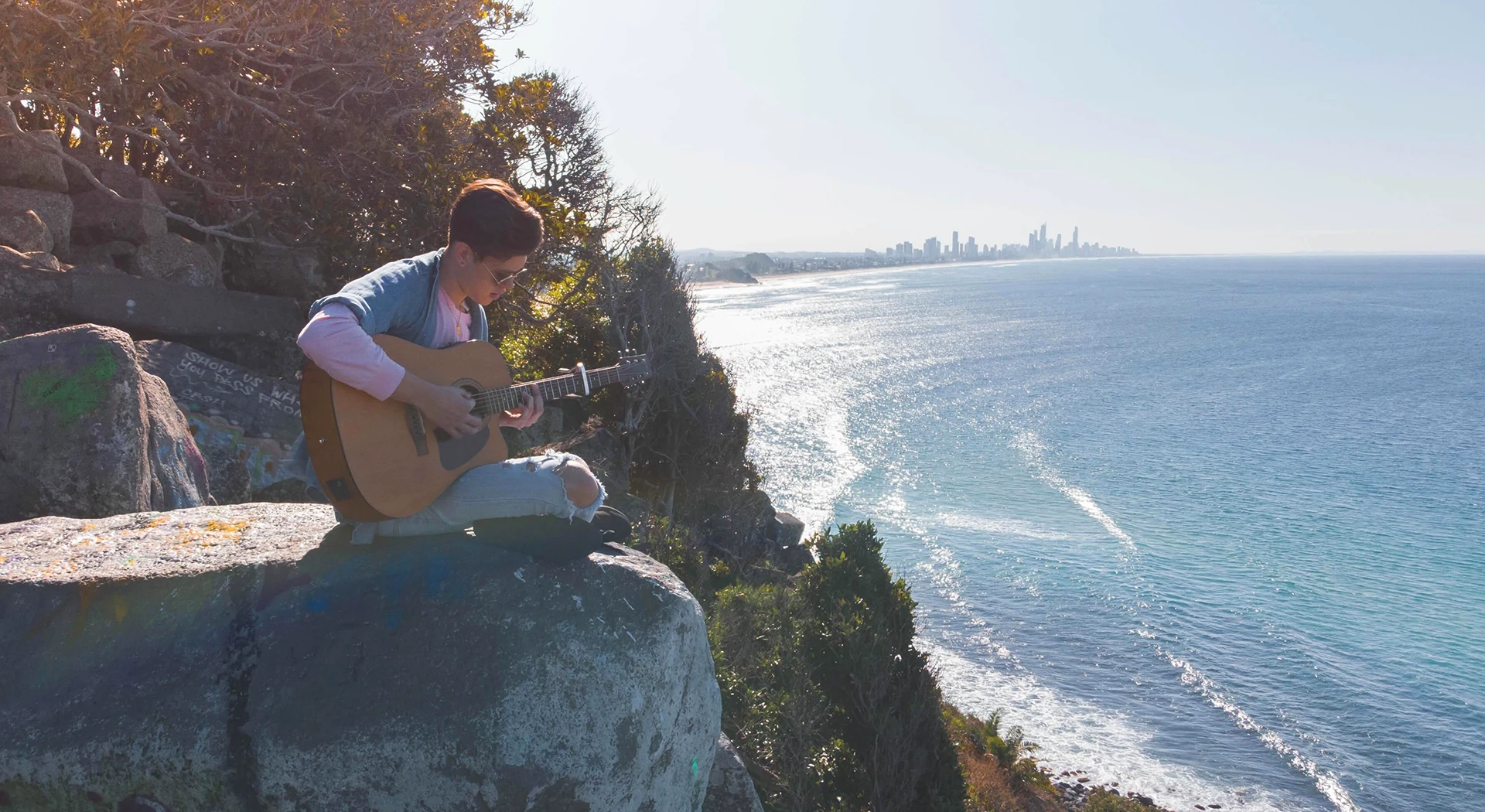 Jacob Muisi sitting with an acoustic guitar above the Gold Coast coastline