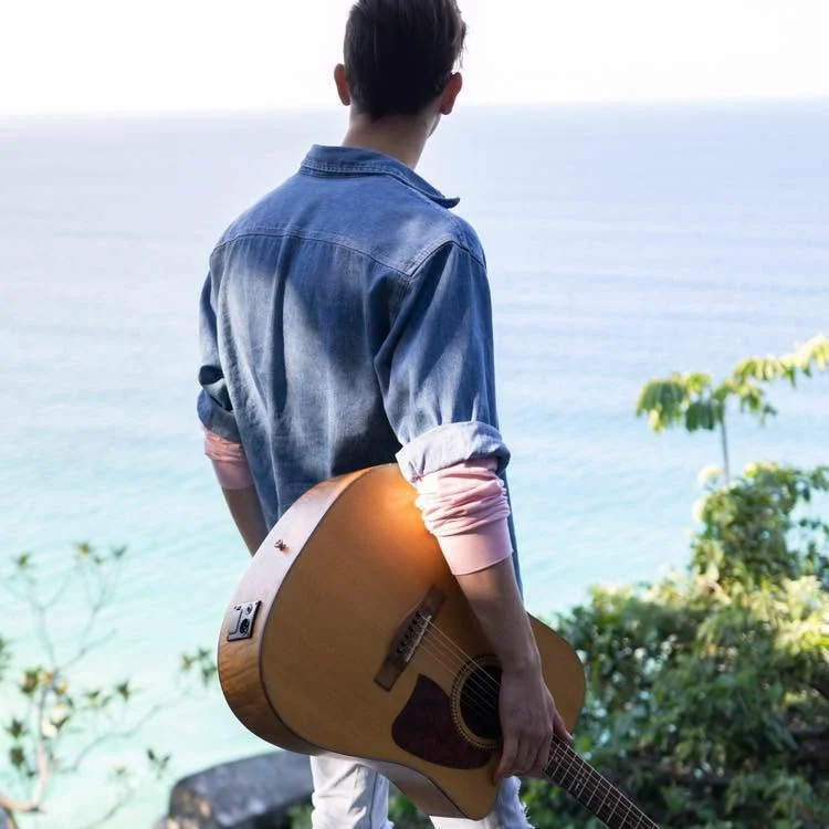 Jacob Muisi holding an acoustic guitar beside the ocean